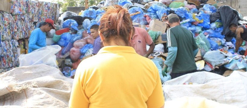 Mulher de costas com blusa amarela olhando para local cheio de lixo